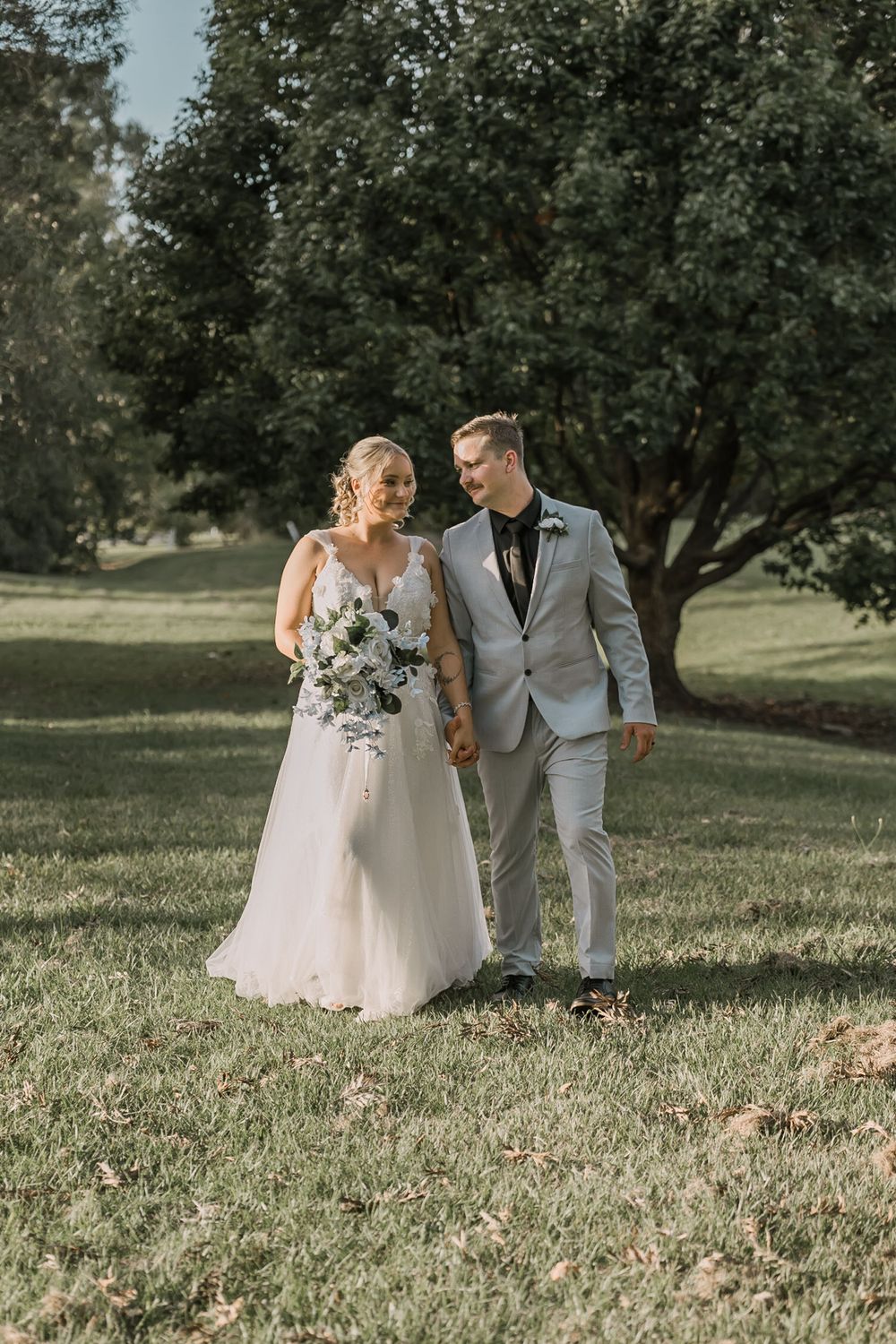 Newlyweds walking together on grass in elegant wedding attire with overhanging trees.