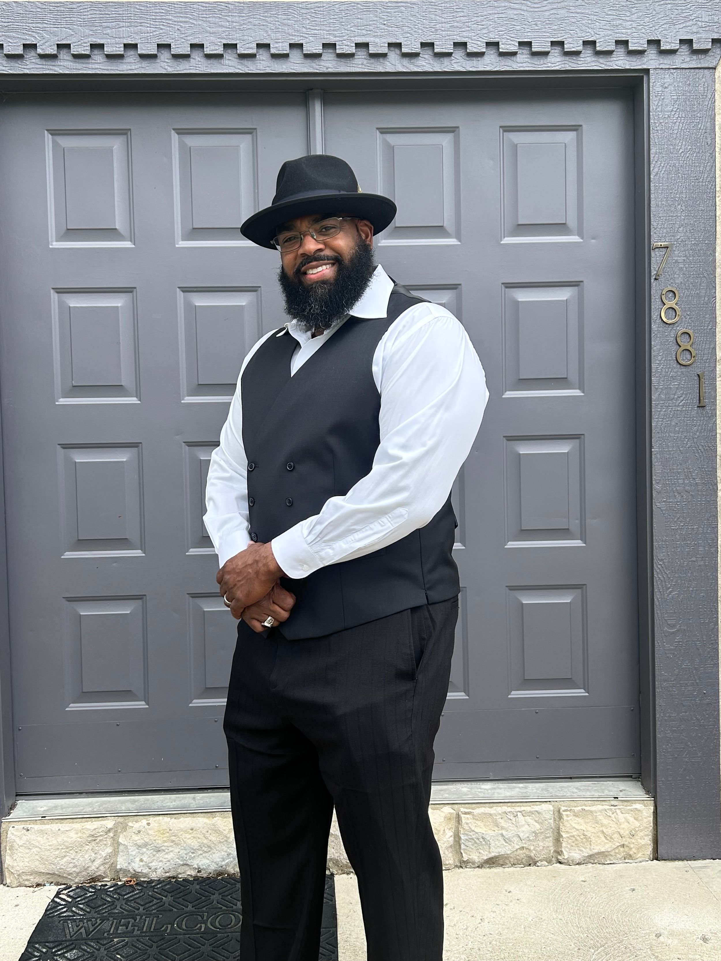 Standing in front of a gray door, someone in classic formal attire with black vest, white shirt, black hat and beard.