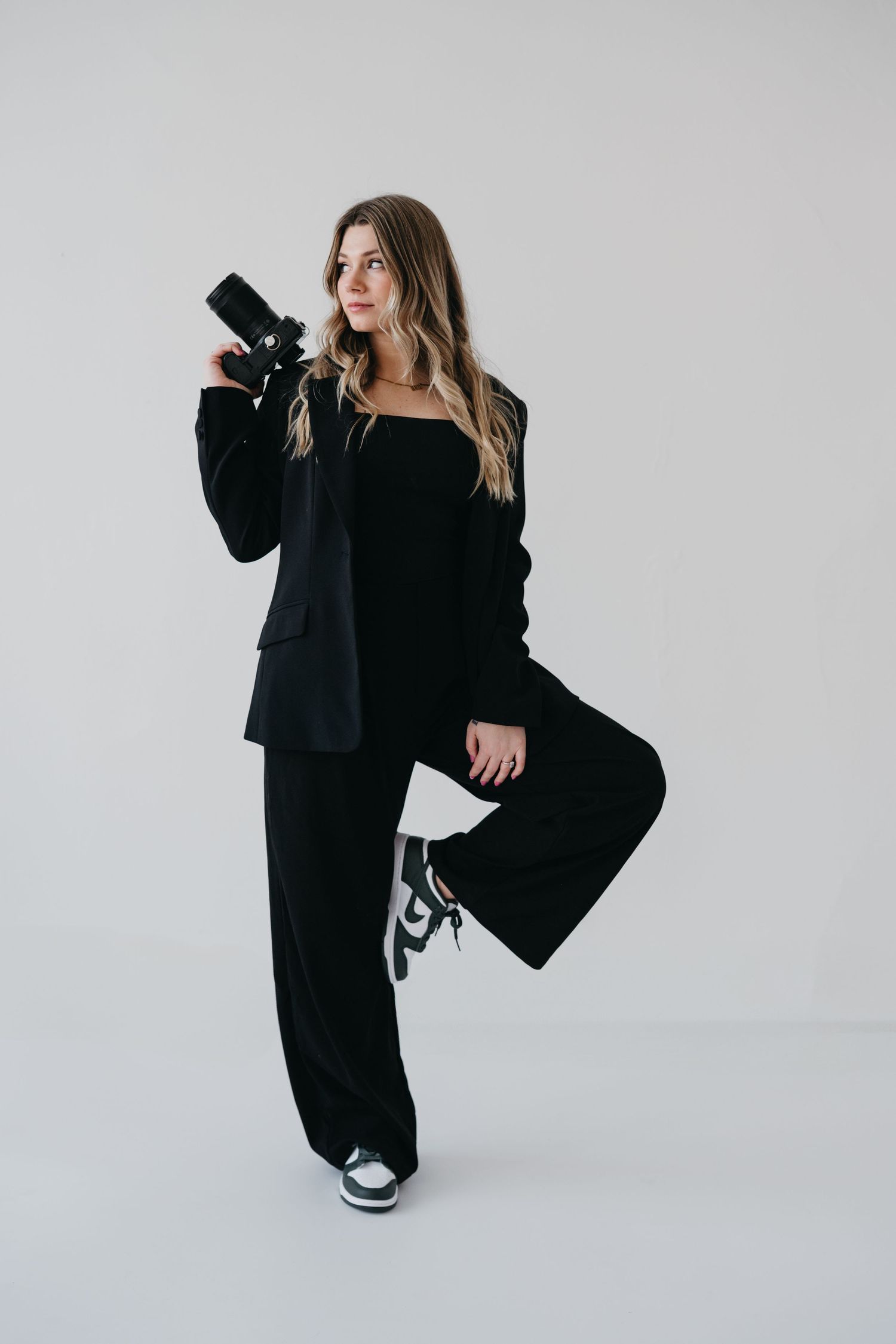 Photographer in all black outfit holding camera against white backdrop while posing with one leg raised.