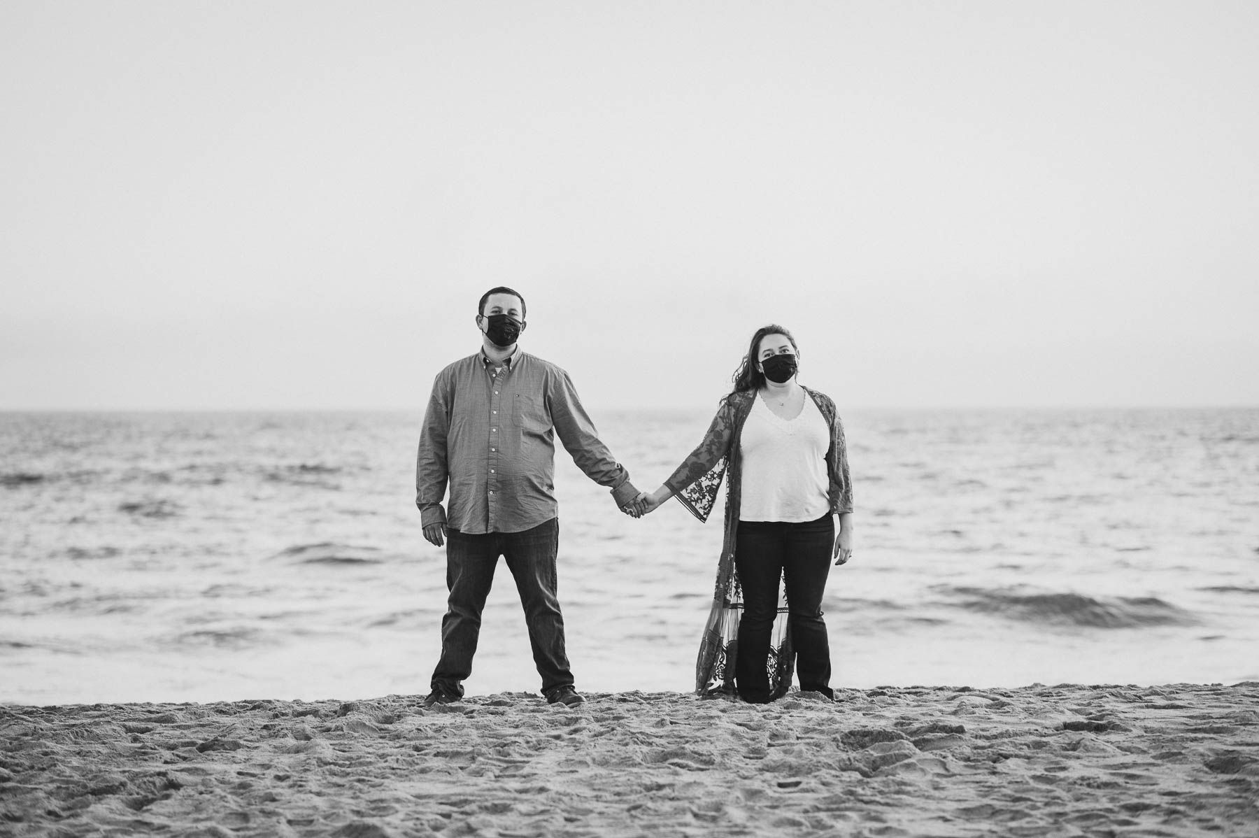Engaged couple posing on the beach while wearing masks in black and white.