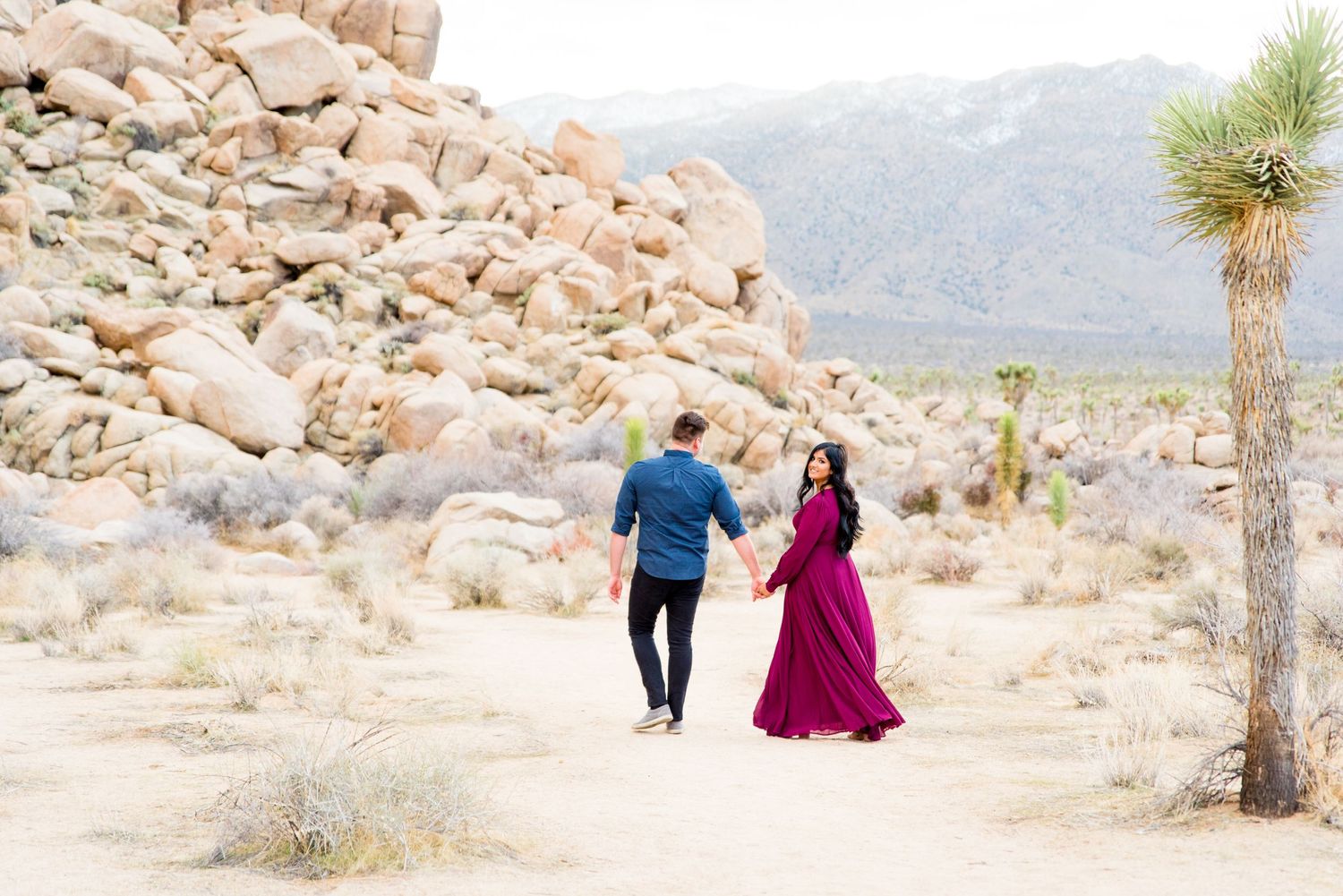 woman in dark red dress looks back as her fiance leads her through Joshua Tree National Park