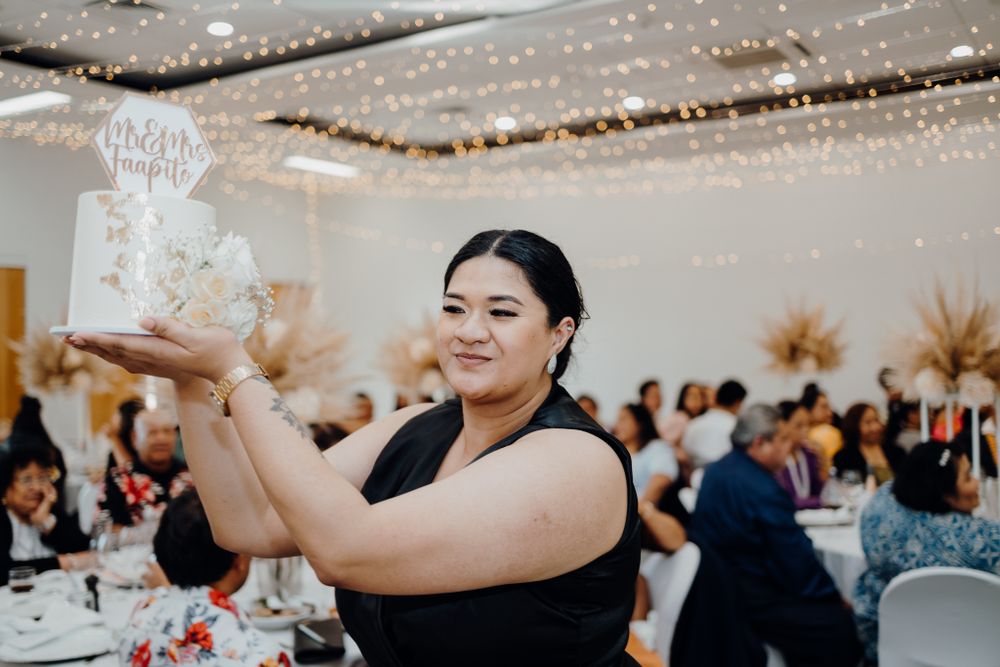 Server in black dress presents elegant white tiered wedding cake at reception.