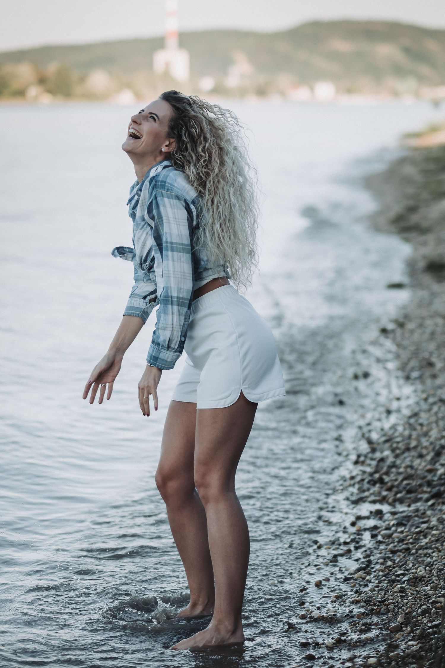 Person with curly hair in plaid shirt enjoying moment at beach with arms spread.