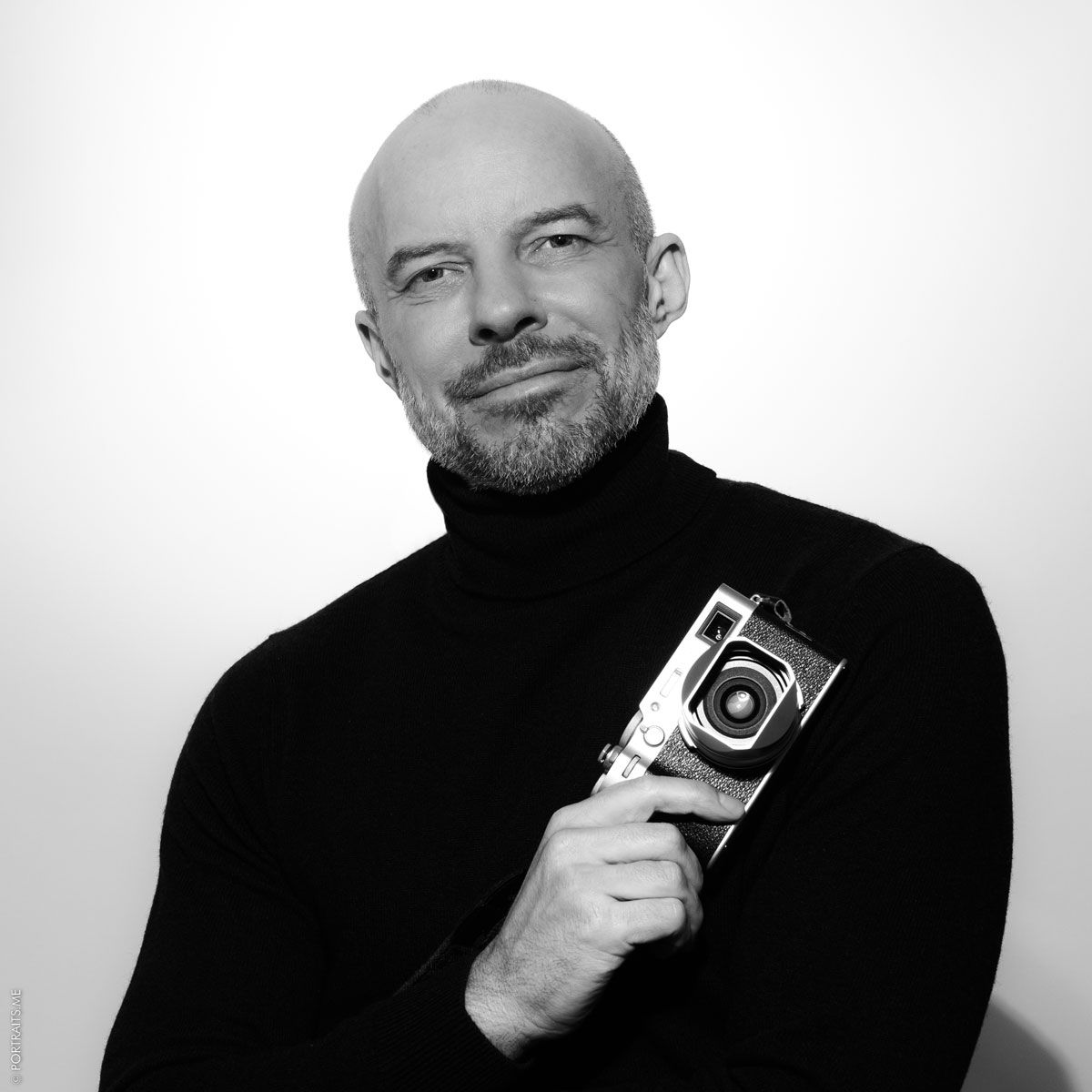 Professional black and white portrait of photographer in black turtleneck holding vintage camera.