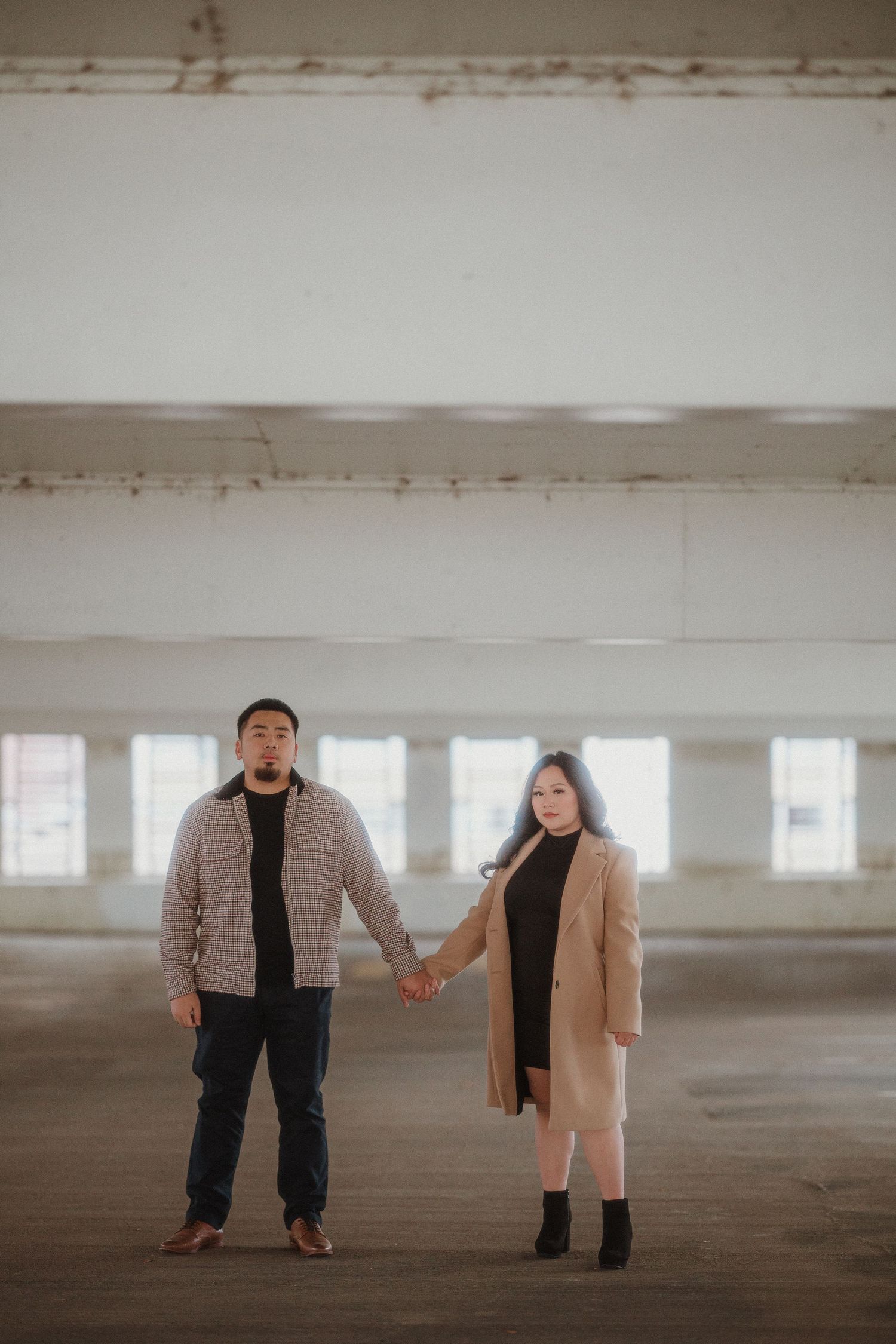 A couple holding hands in an empty parking garage with a minimalist industrial backdrop.
