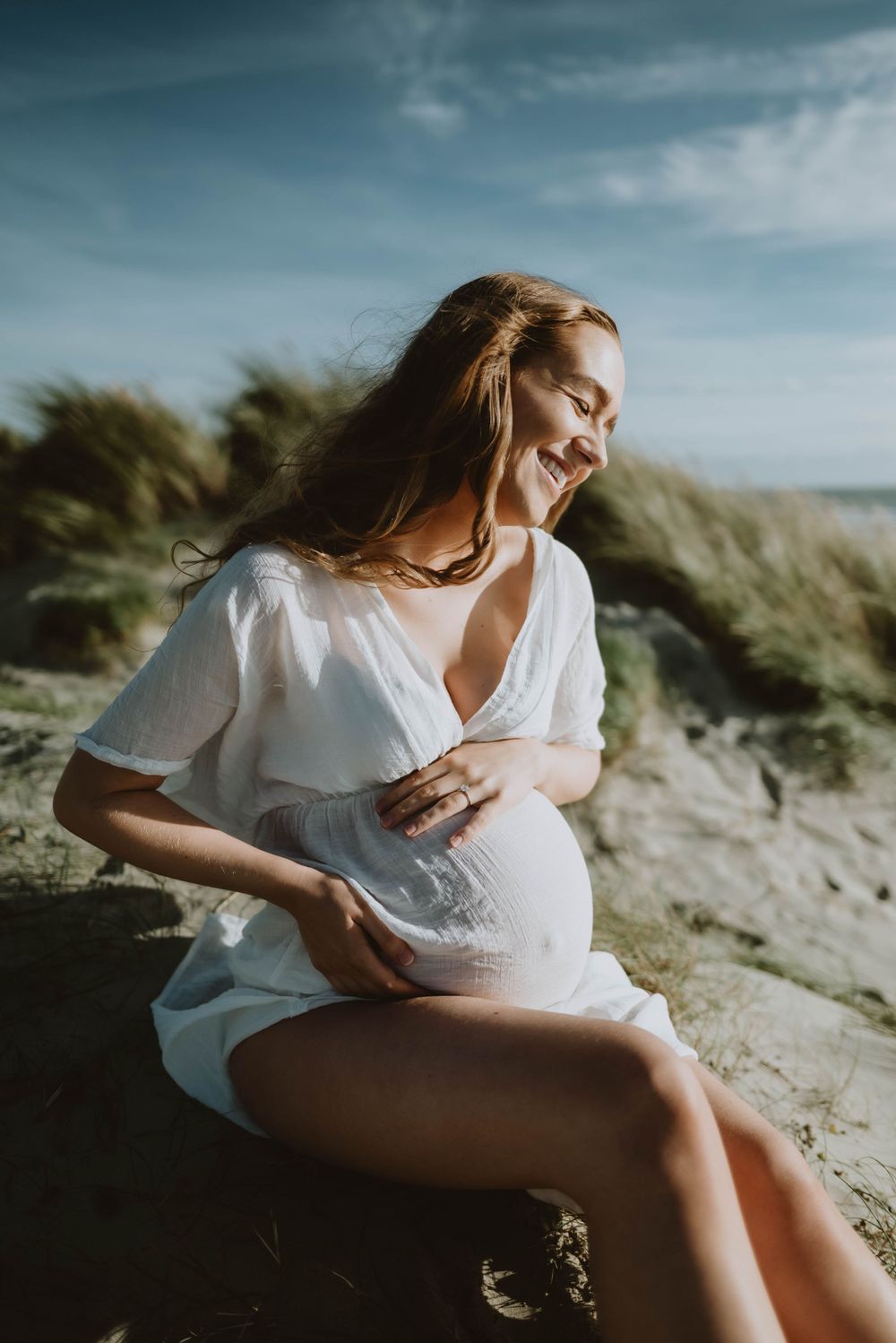 An expectant mother in white dress sits on coastal rocks during golden hour.