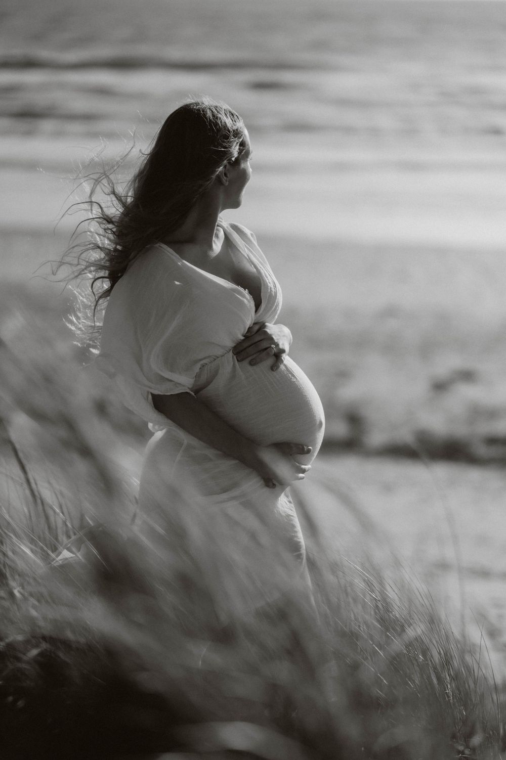 A dreamy black and white maternity photo showing a pregnant woman in a white dress standing in tall grass.