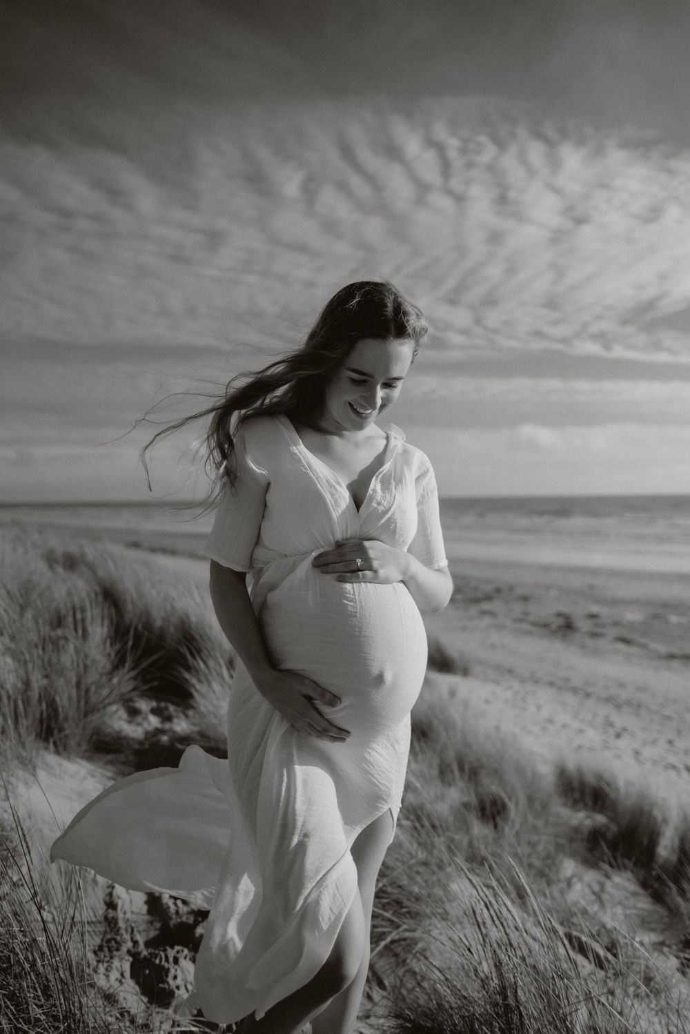 A pregnant woman in a flowing white dress stands on a beach under dramatic cloudy skies in black and white photography.