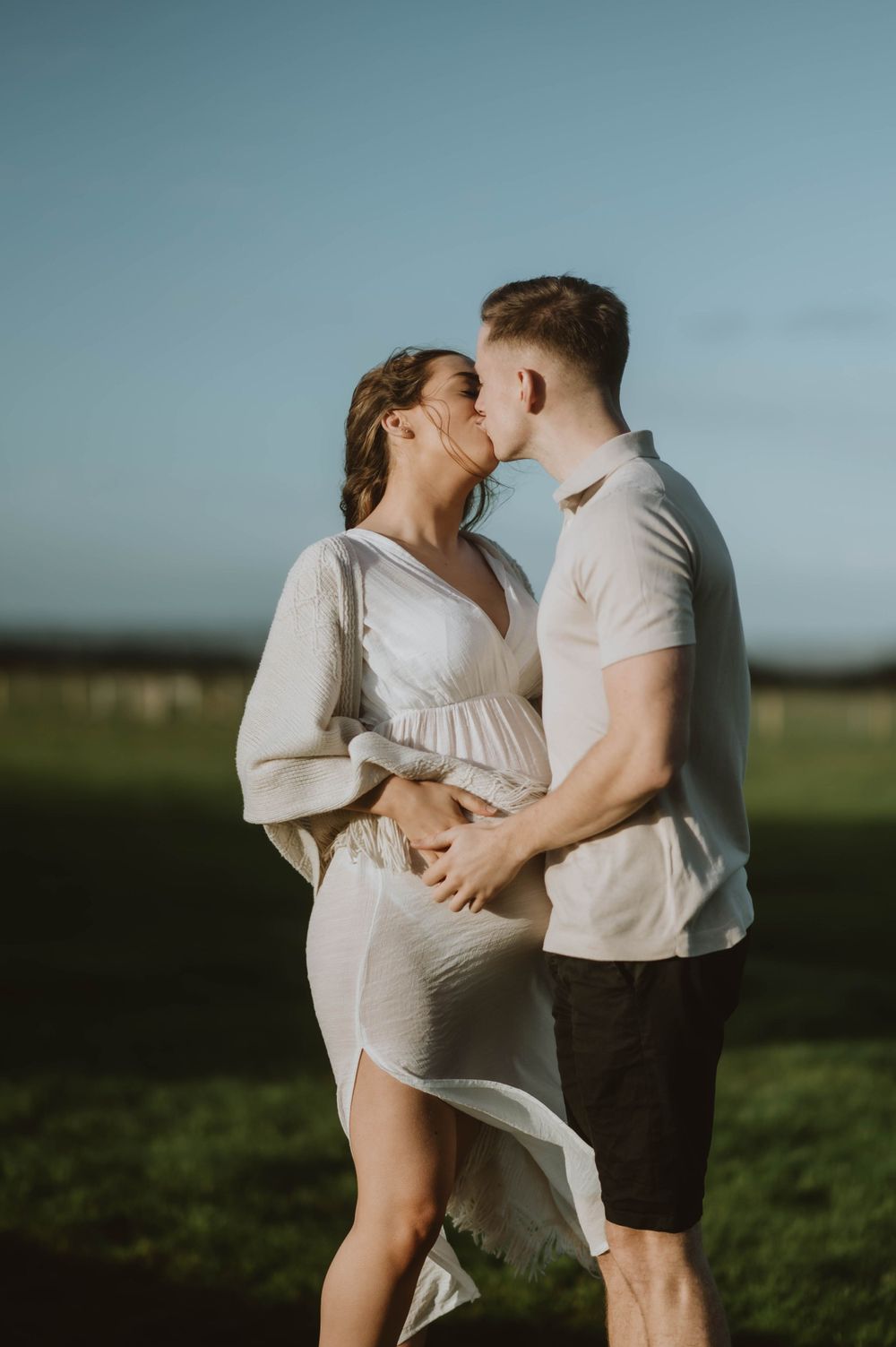 A romantic couple in white clothing share a tender moment together in a sunlit field against a blue sky.