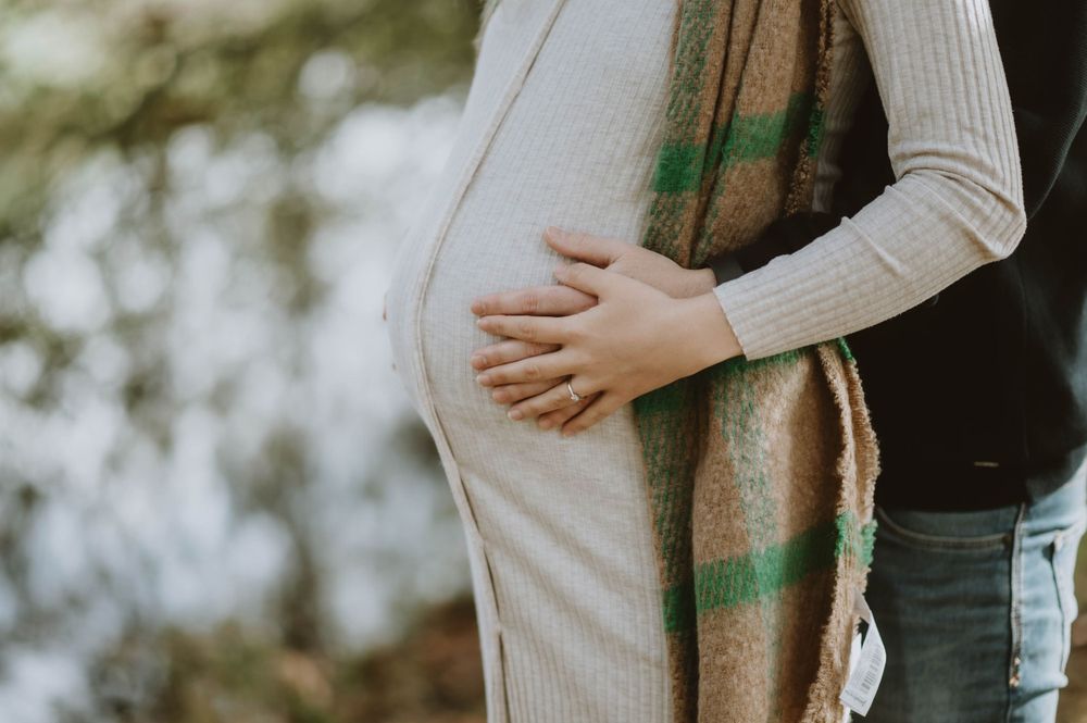 A close-up shot of a pregnant belly being cradled by hands in a beige sweater against a soft bokeh background.