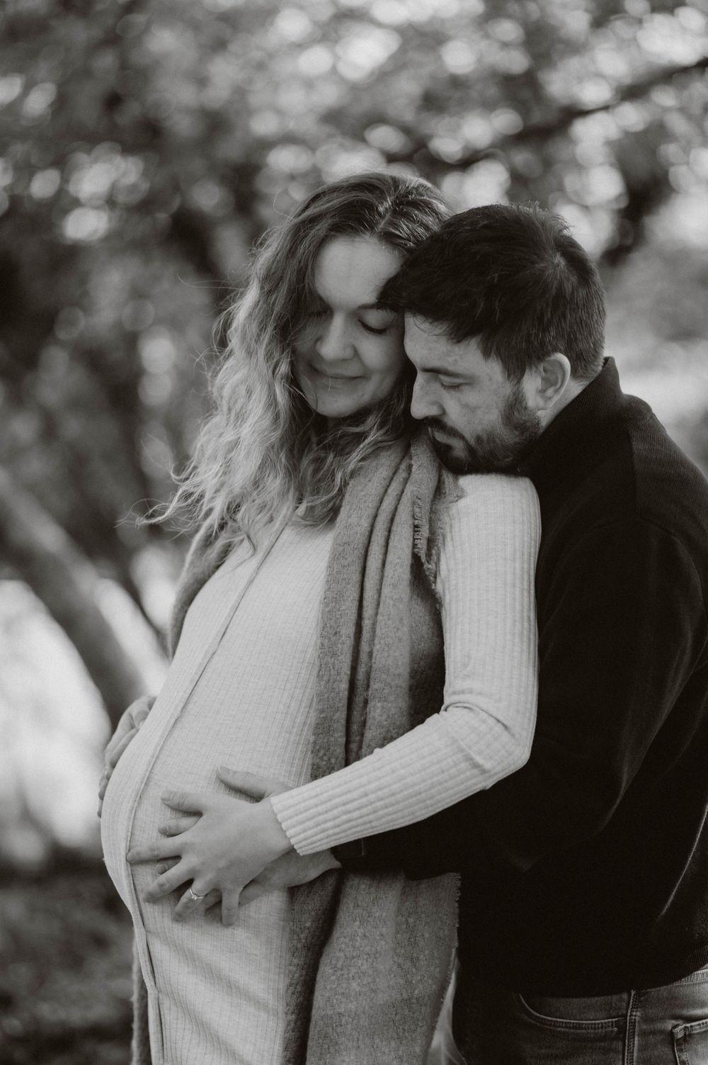 A tender black and white maternity photo shows a couple embracing outdoors with soft bokeh in the background.