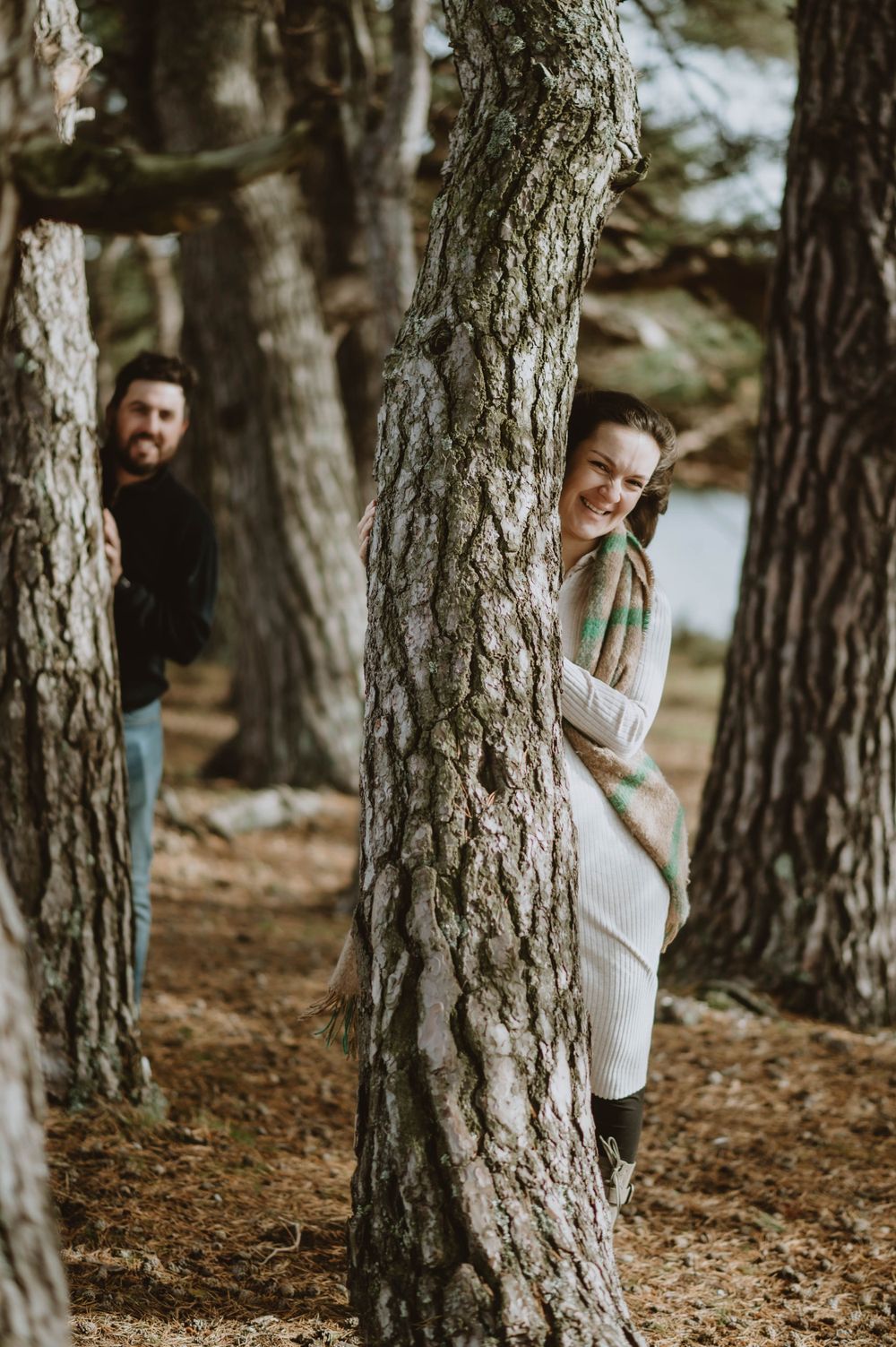 Playful couple peek out from behind large tree trunks in a pine forest on a sunny afternoon.