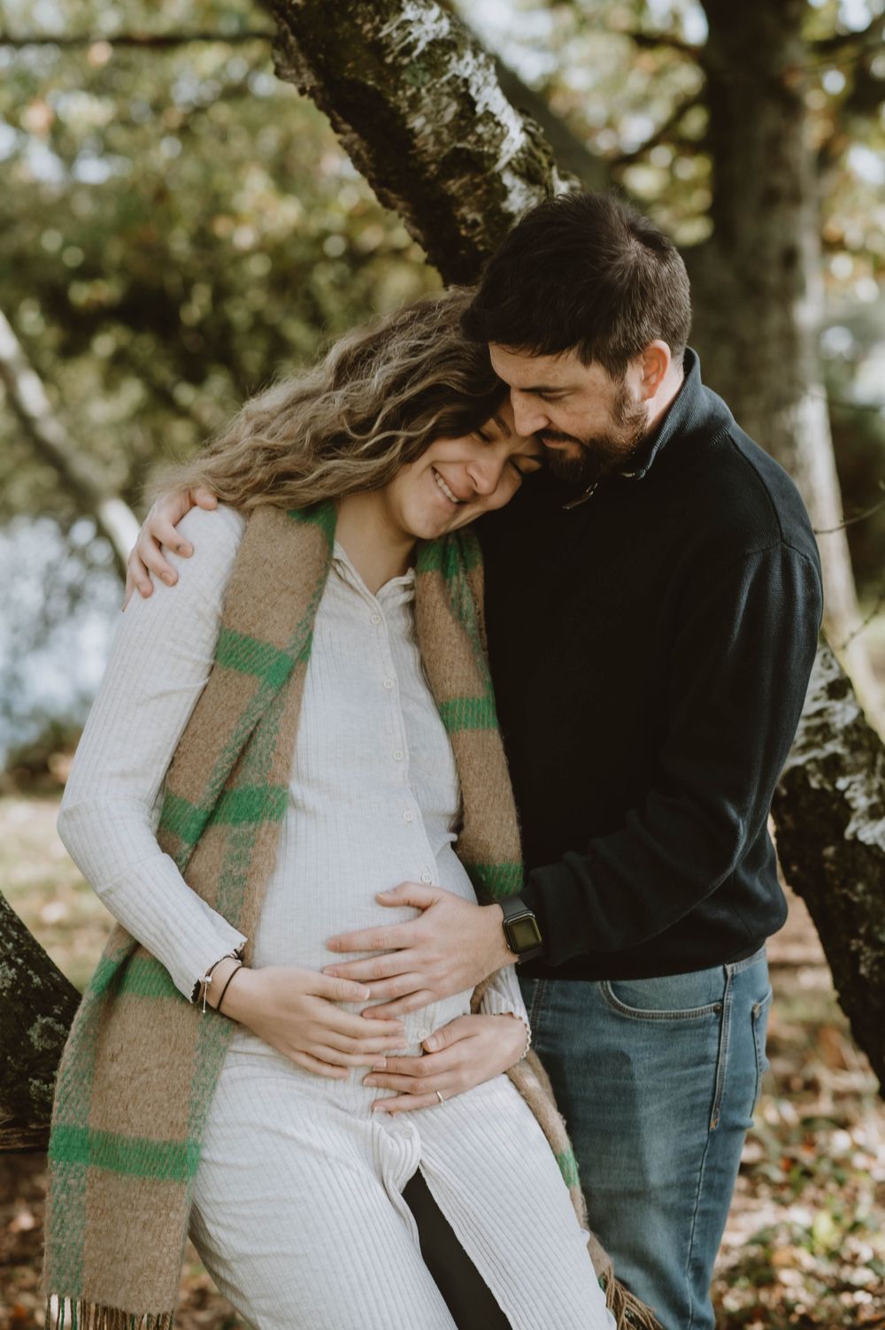 A couple shares an intimate moment outdoors during a maternity photo shoot surrounded by autumn trees.