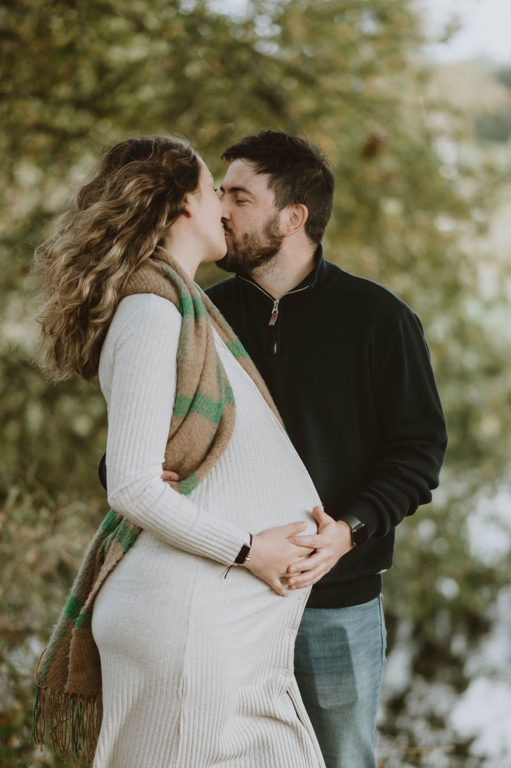 A romantic maternity photo of a couple embracing outdoors in autumn with the woman wearing a cream sweater dress.