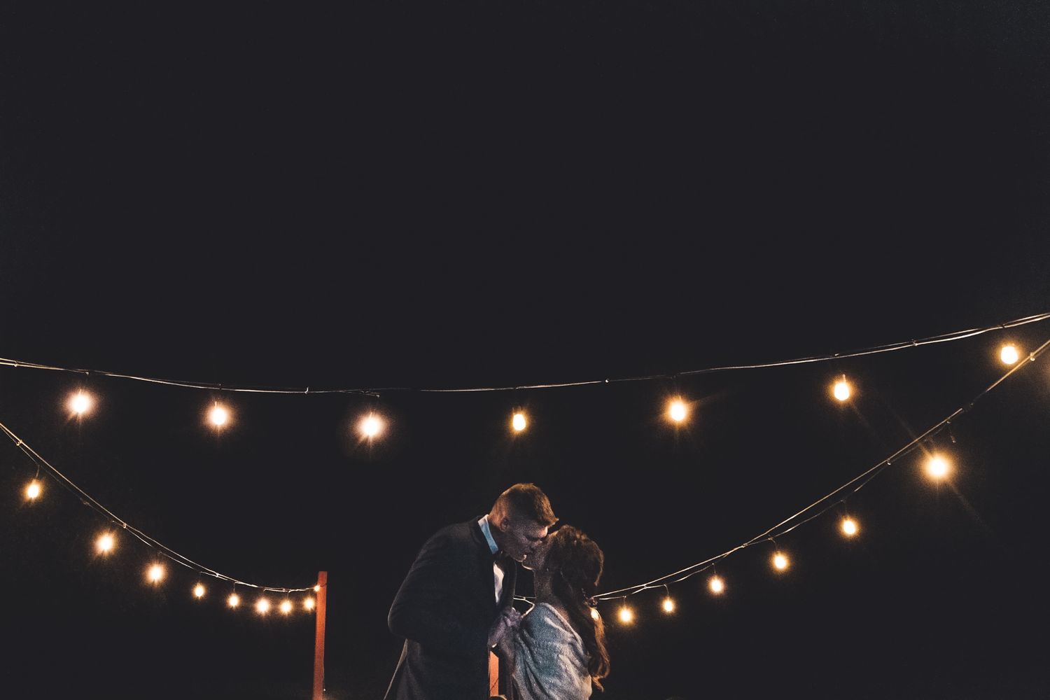 A romantic silhouette under string lights at night creating an intimate wedding moment against a dark sky.