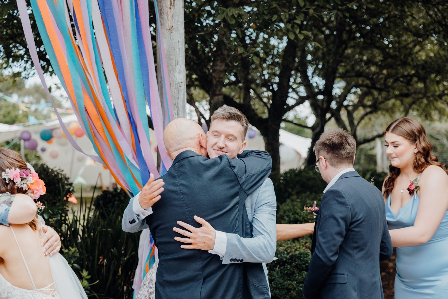 Wedding guests sharing warm embraces during outdoor ceremony with colorful ribbon decor.