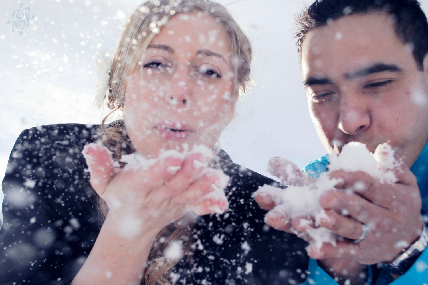 Two people playfully blowing snow at each other in a winter scene against a bright background.