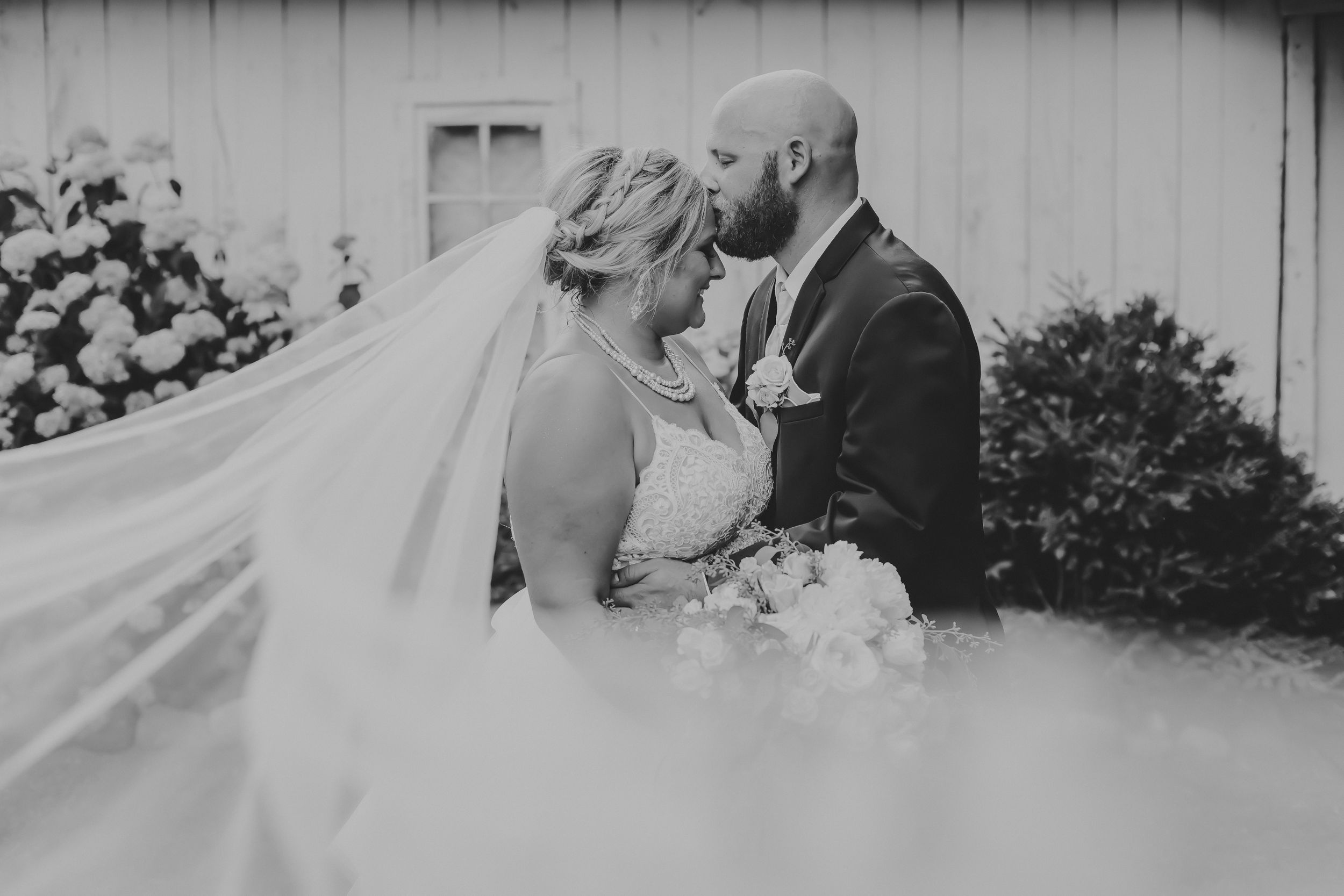Black and white photo of groom kissing bride's forehead. Her cathedral veil blows toward the camera.