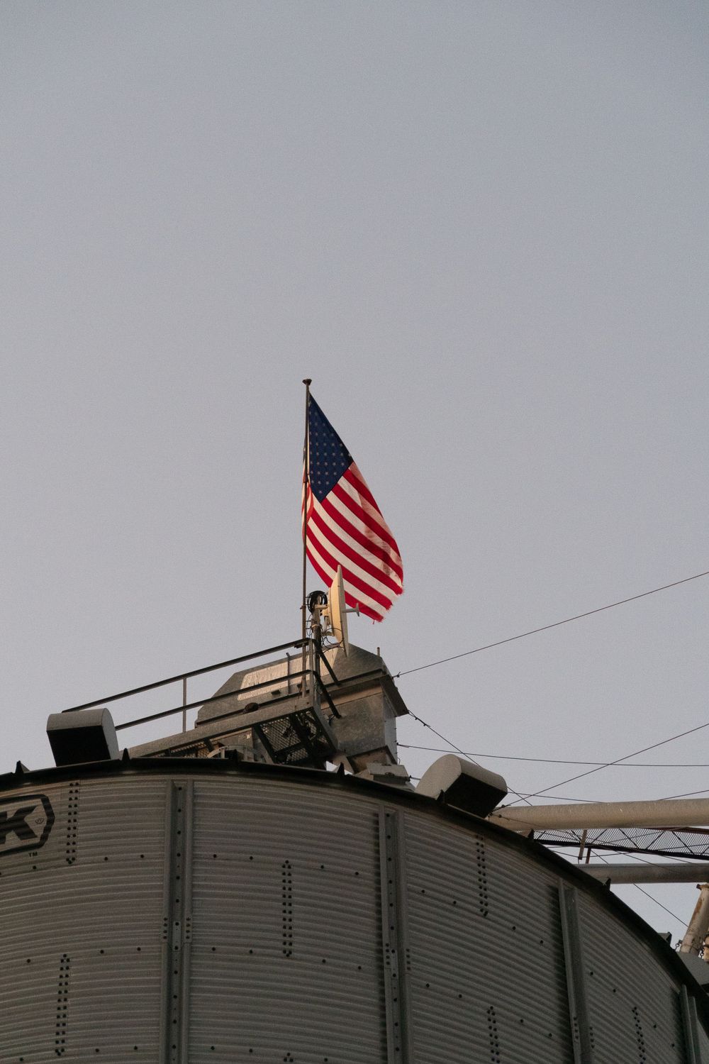 American flag waves atop metal grain silo against grey sky.
