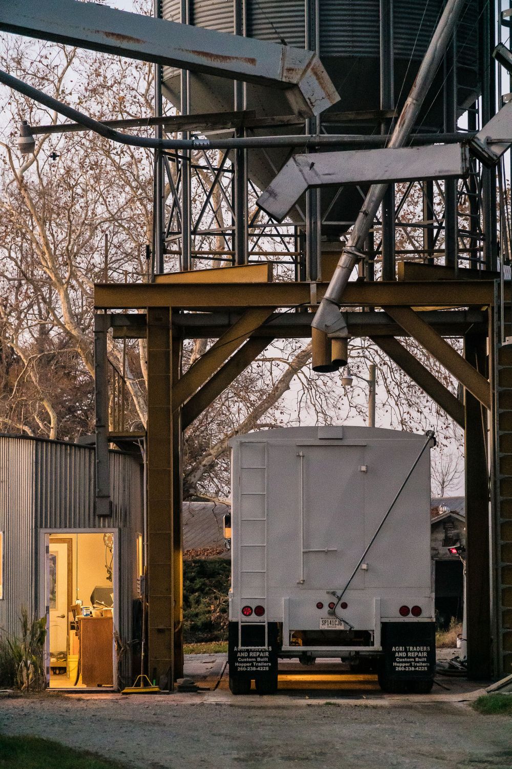 Semi truck trailer parked under grain loading structure at dusk.
