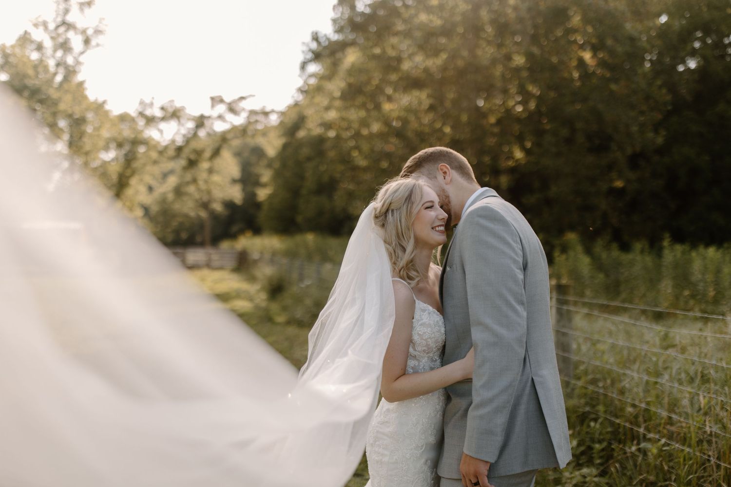 Couple embracing in wedding attire during sunset in rustic outdoor setting.