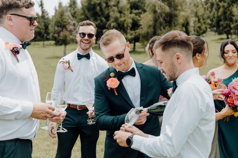 Wedding party members in suits share laughs and drinks on a sunny outdoor lawn.