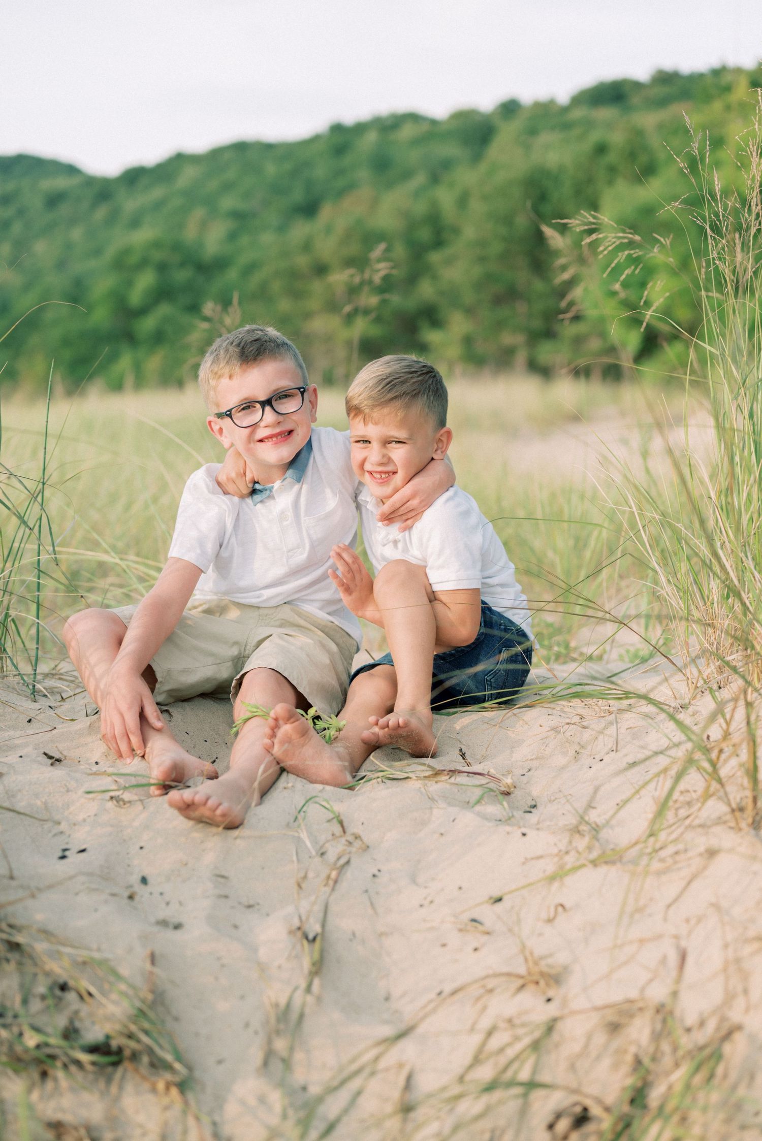 Two children sit together on sandy dunes, sharing laughs in casual summer attire.
