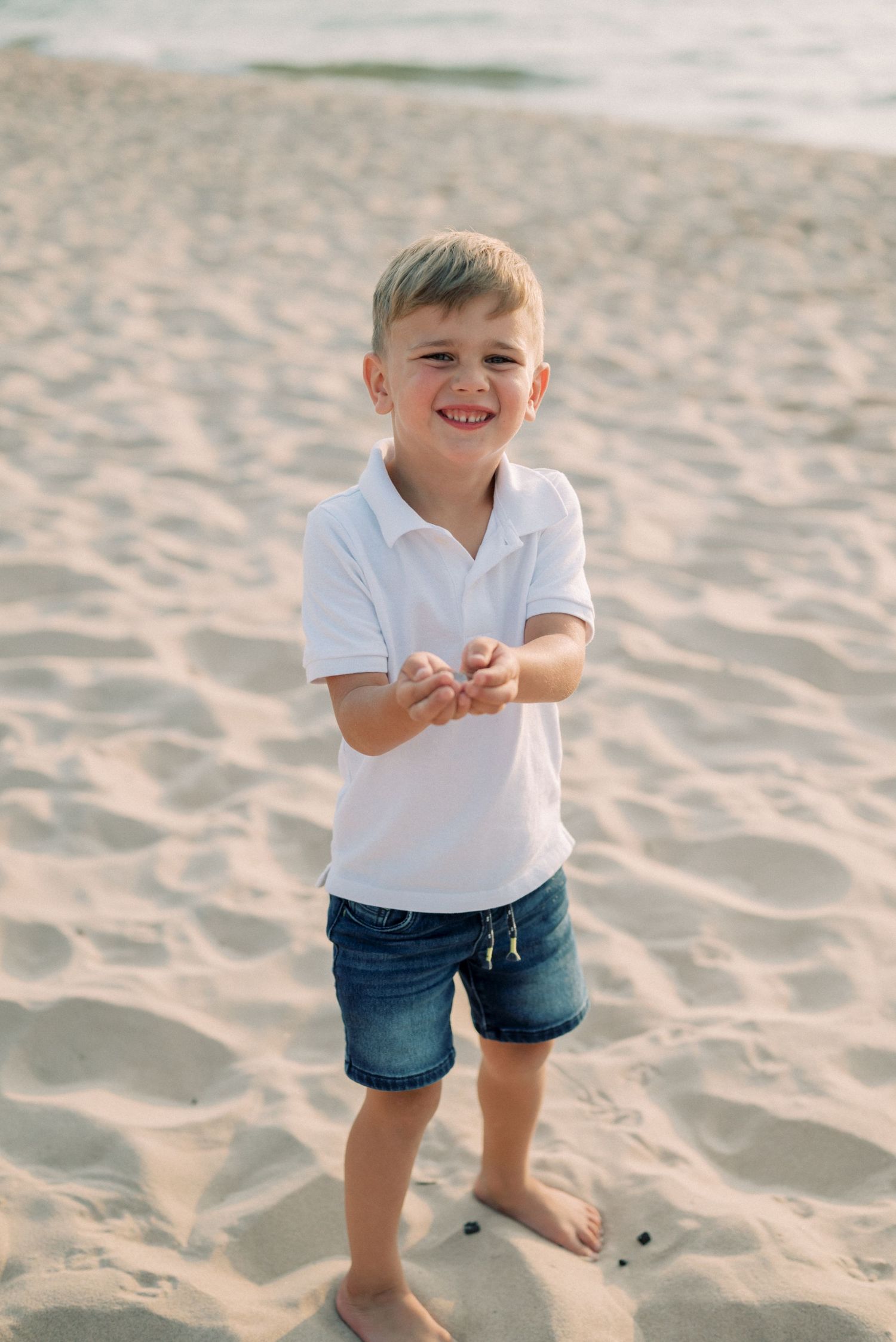 Young child in a white polo and shorts laughing while playing on a sandy beach.