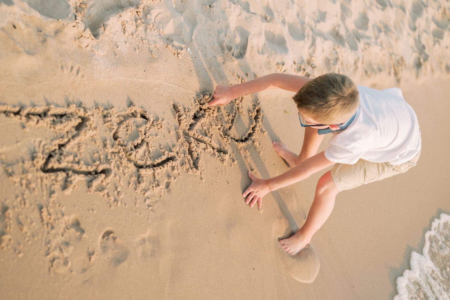 Child writing in the wet sand at beach edge as waves approach.