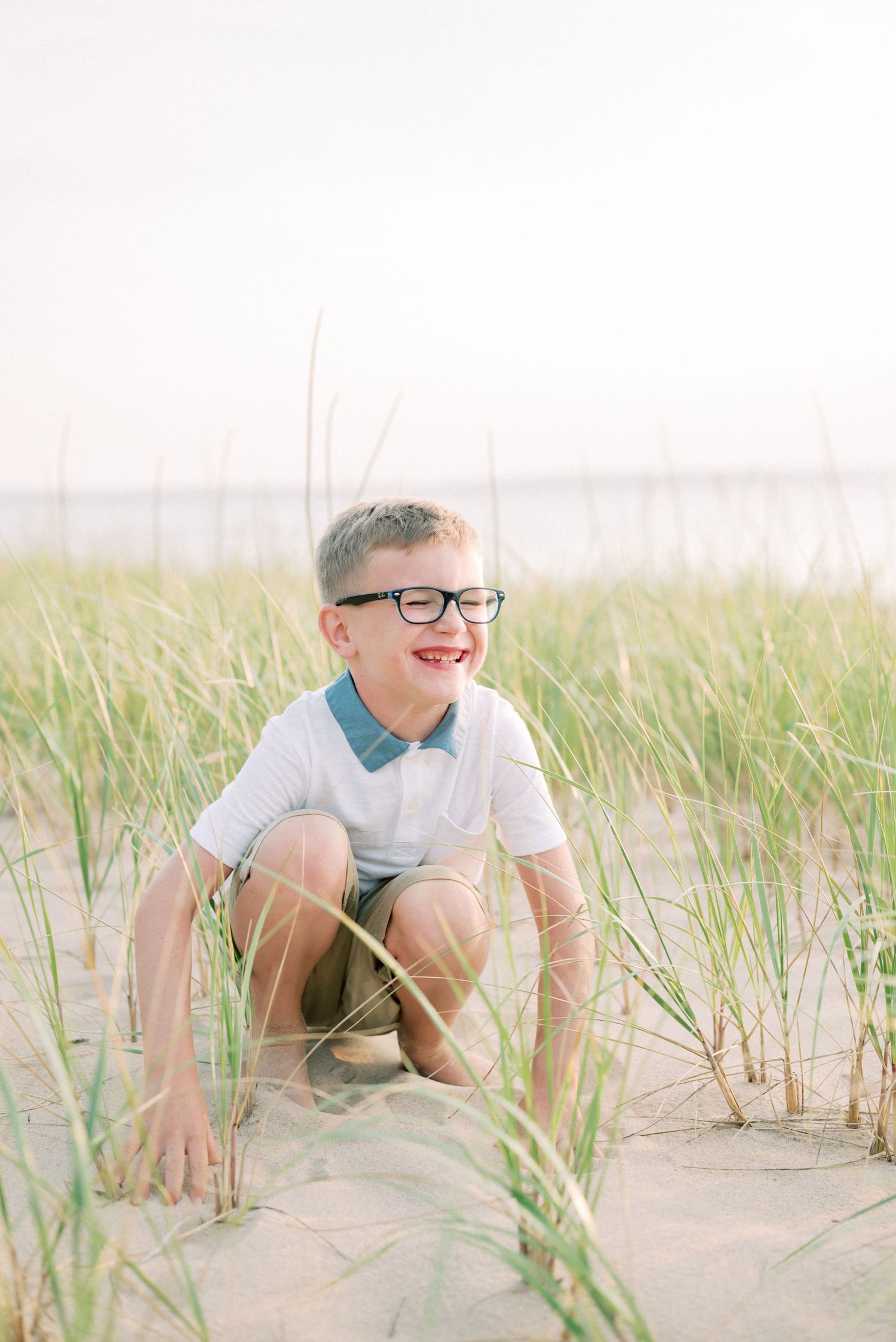 Child crouching among beach grass with a bright smile and glasses.