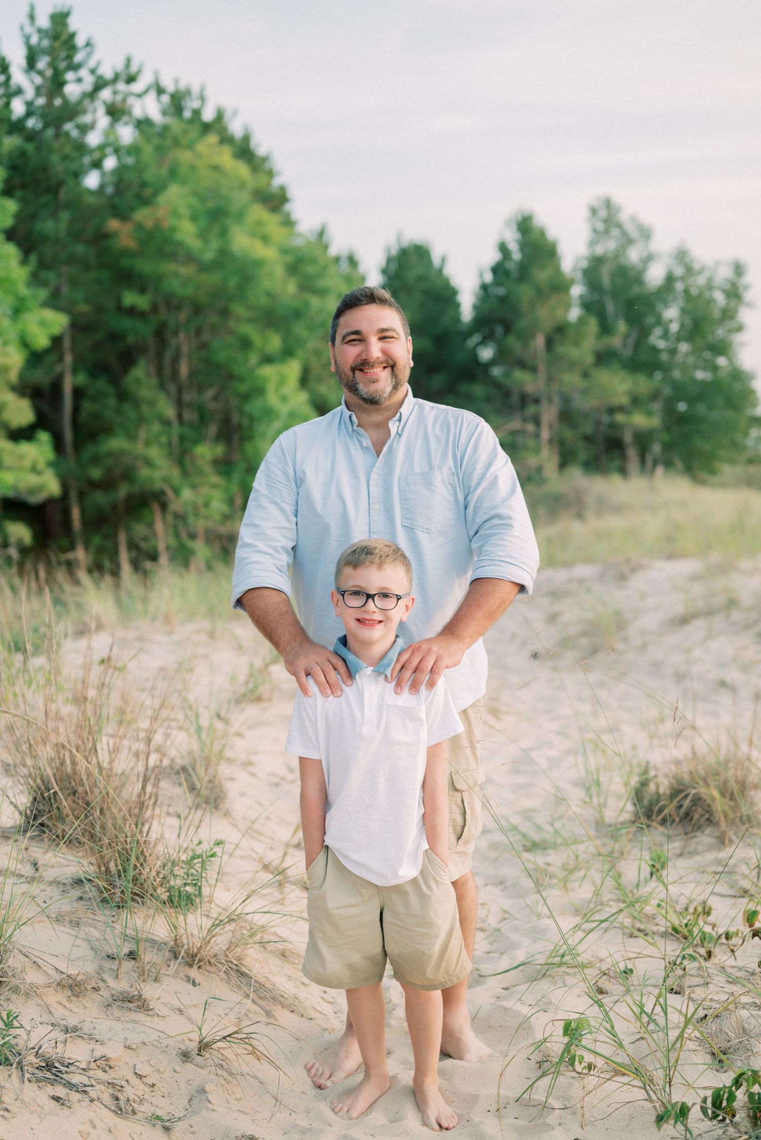 A casual beachside portrait on sandy dunes with pine trees in the background.
