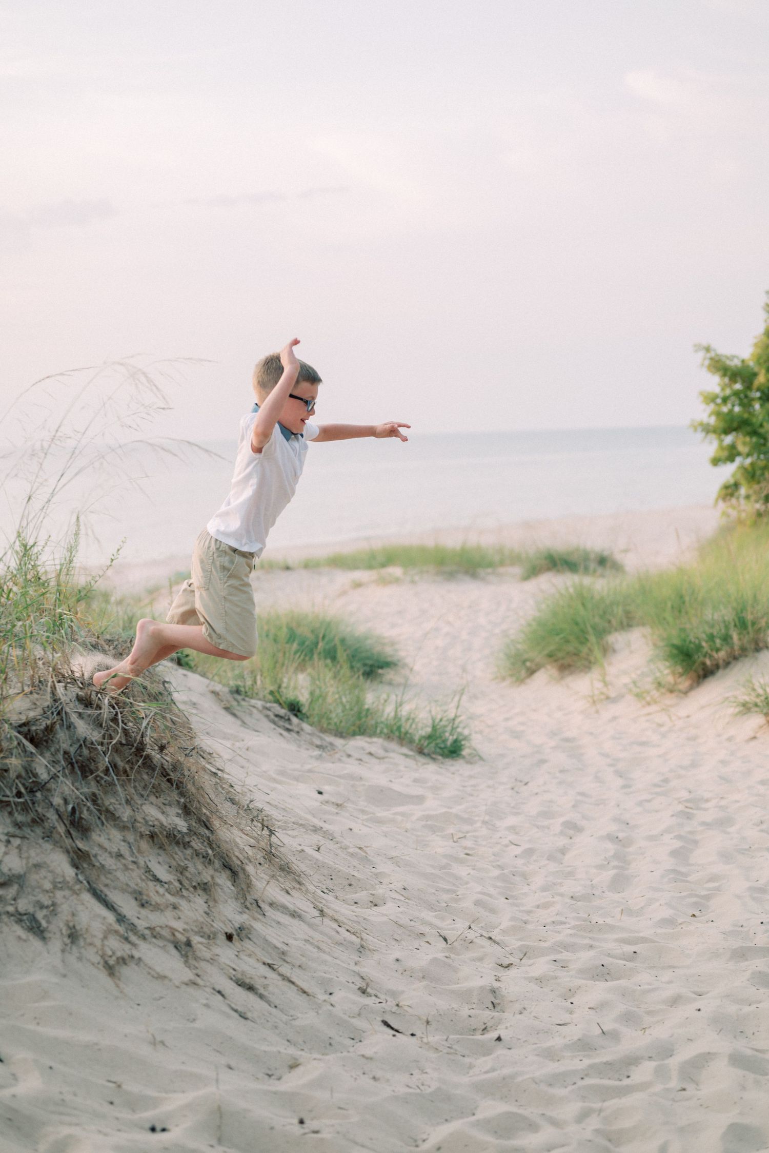 Child leaping from sandy dune with joy against coastal landscape backdrop.
