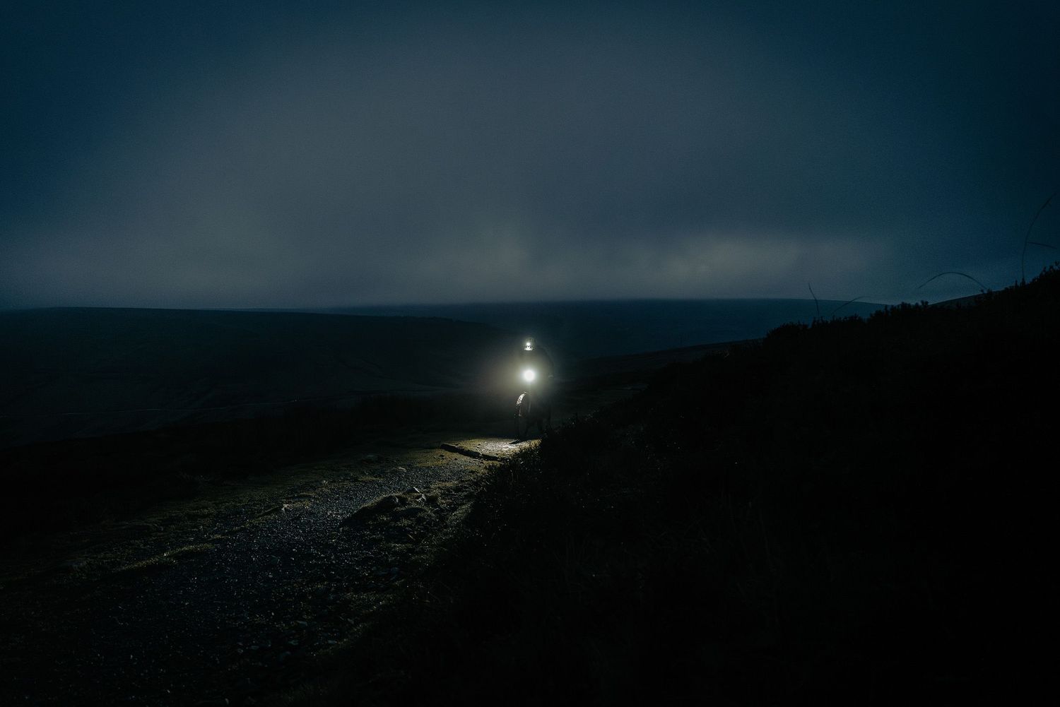 A bright headlamp illuminates a dark coastal path at night with moody stormy skies overhead.