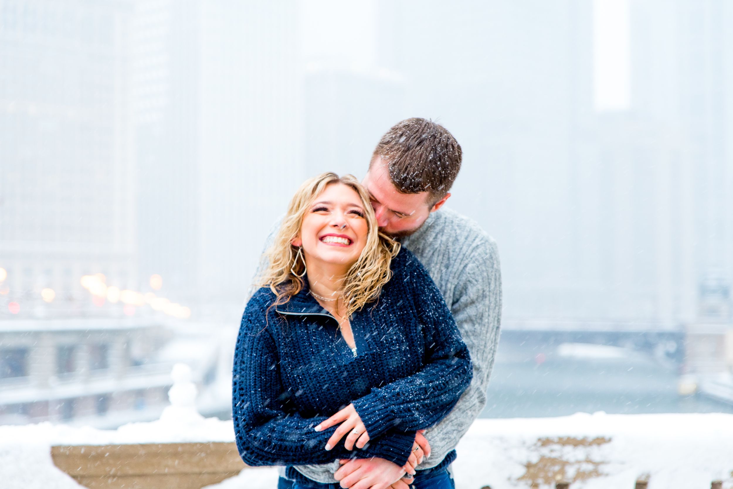 man in gray sweater hugging and kissing fiance in navy sweater at Wrigley Building for Chicago winter engagement picture