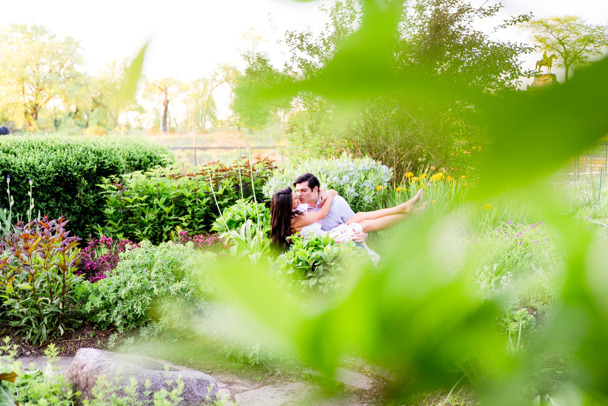 woman sitting on fiance's lap in Lincoln Park as they kiss for engagement pictures in the garden