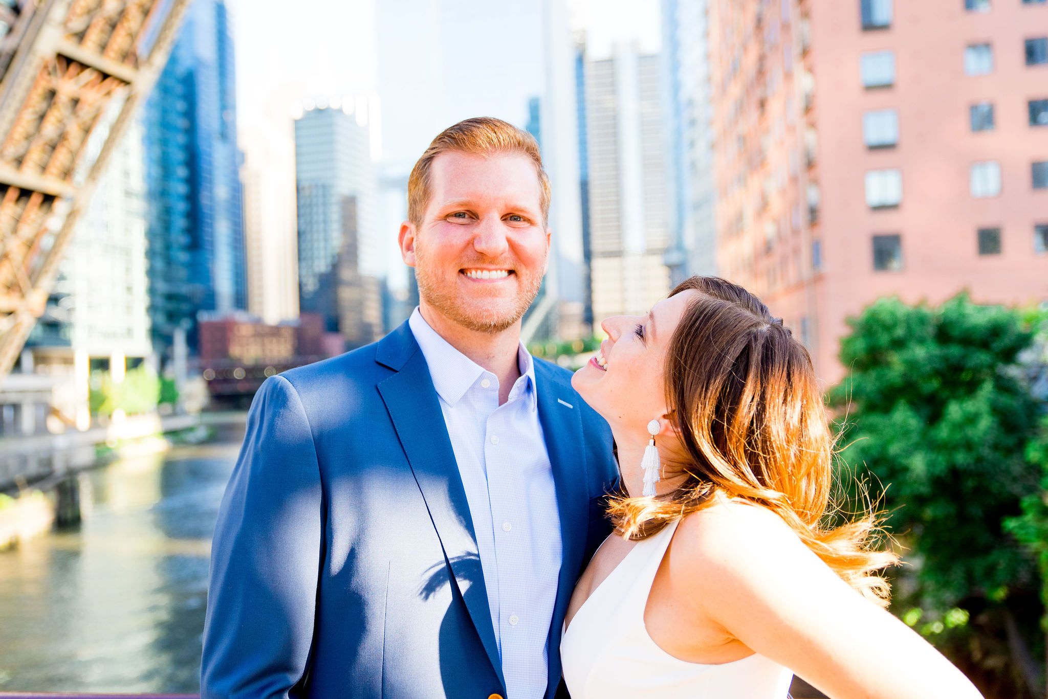 woman in white dress smiling at man in navy suit in front of Chicago River at Kinzie St Bridge engagement pictures
