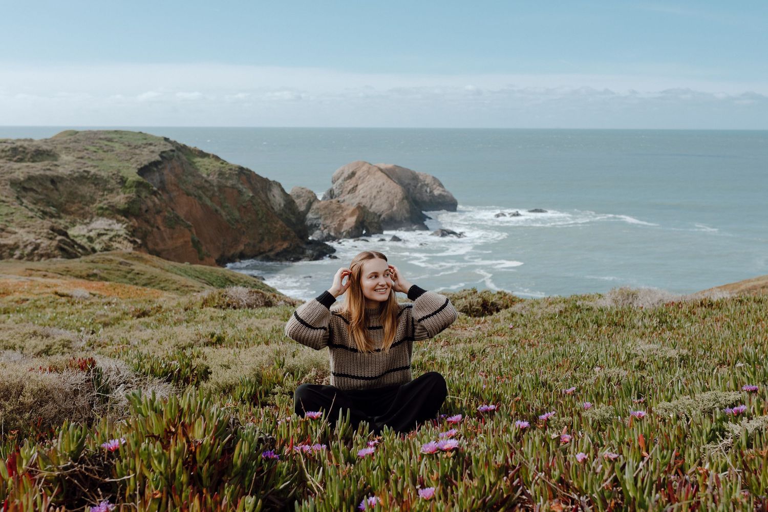 Happy person posing with arms raised among coastal wildflowers with ocean cliffs in background.