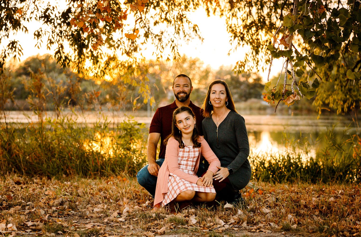 family of 3 squatting down psoing for liz the photographer during a fall session in colorado in front of a lake during sunset golden hour