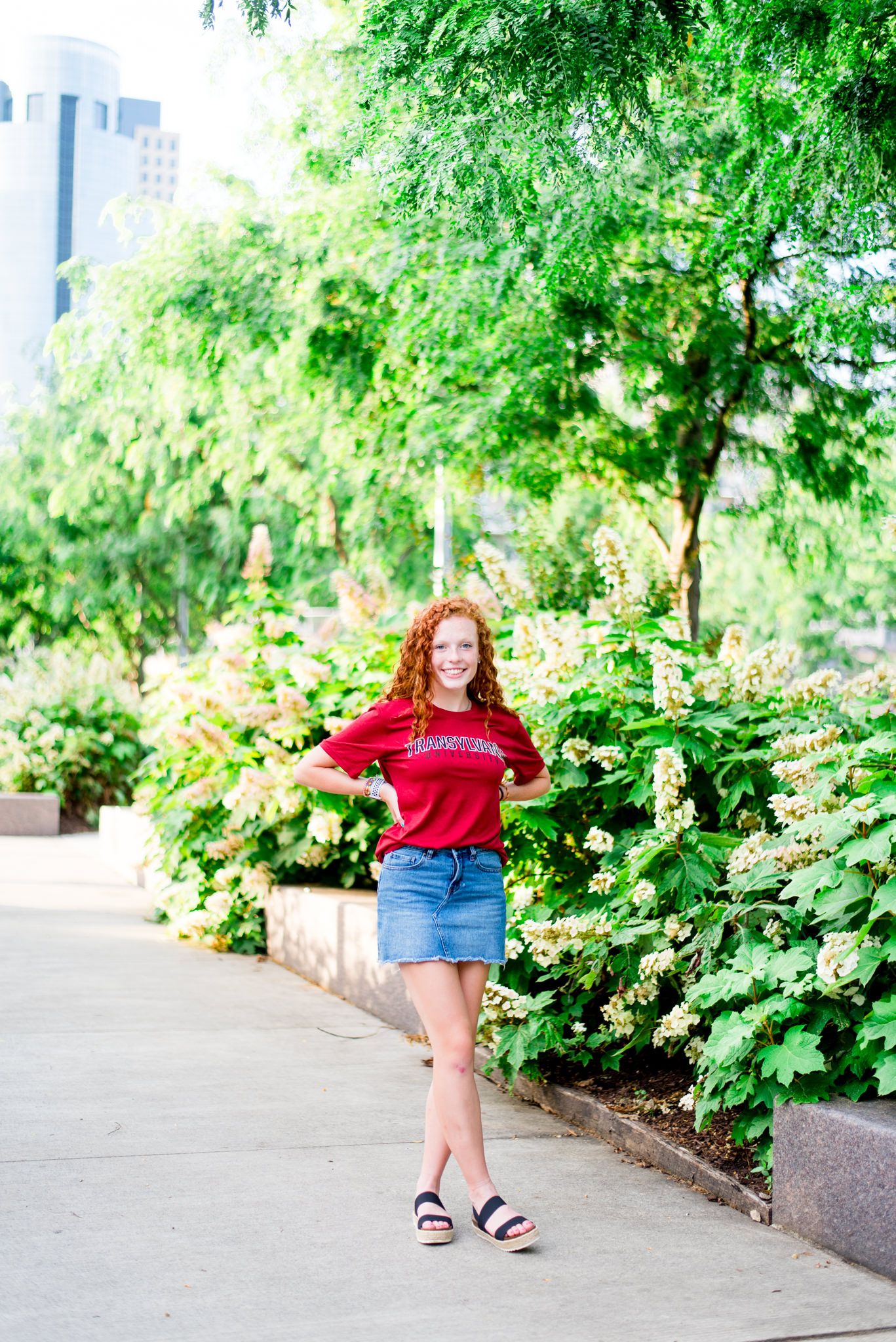 young woman with red curly hair in red Transylvania shirt and denim skirt walking through Smale Park hydrangeas