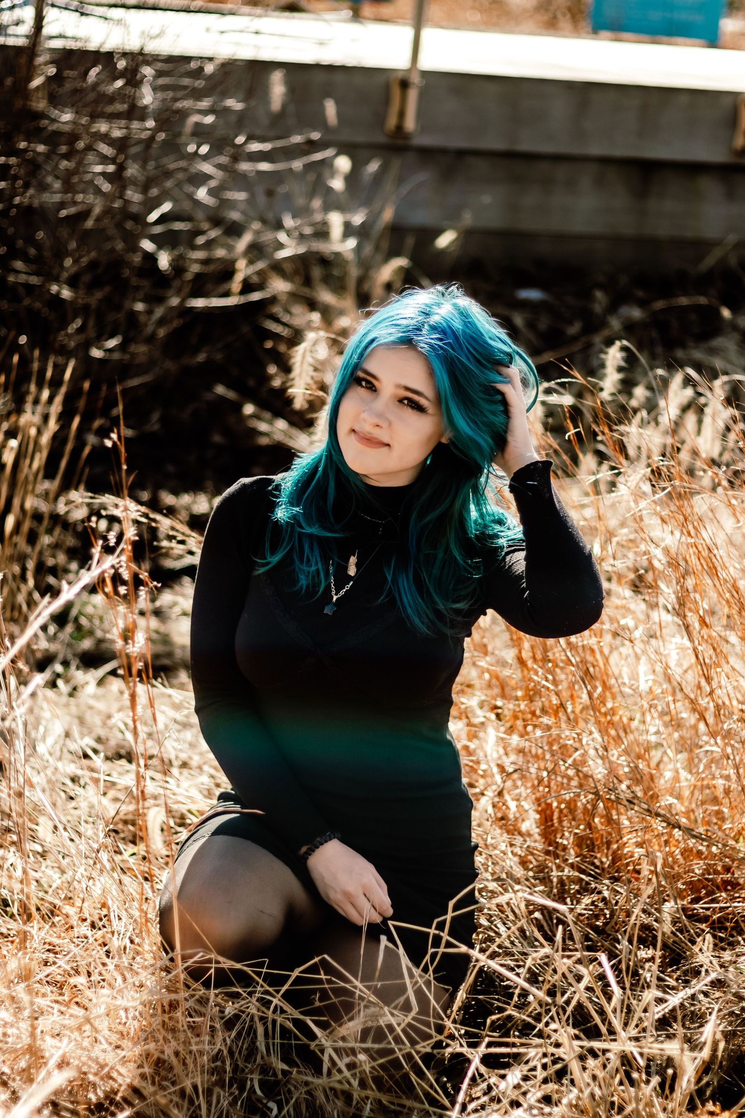 Portrait in tall grass featuring vibrant teal hair and black outfit with golden sunlit backdrop.
