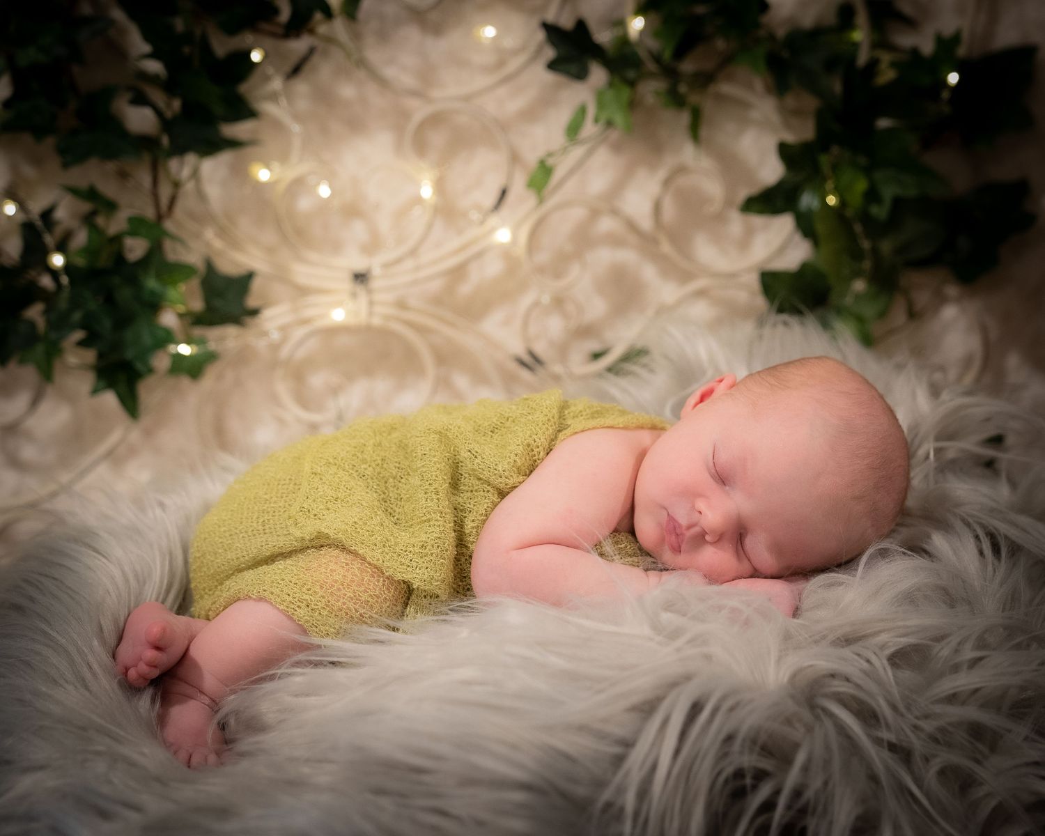 Sleeping infant in yellow wrap lies on soft white fur blanket with greenery backdrop.