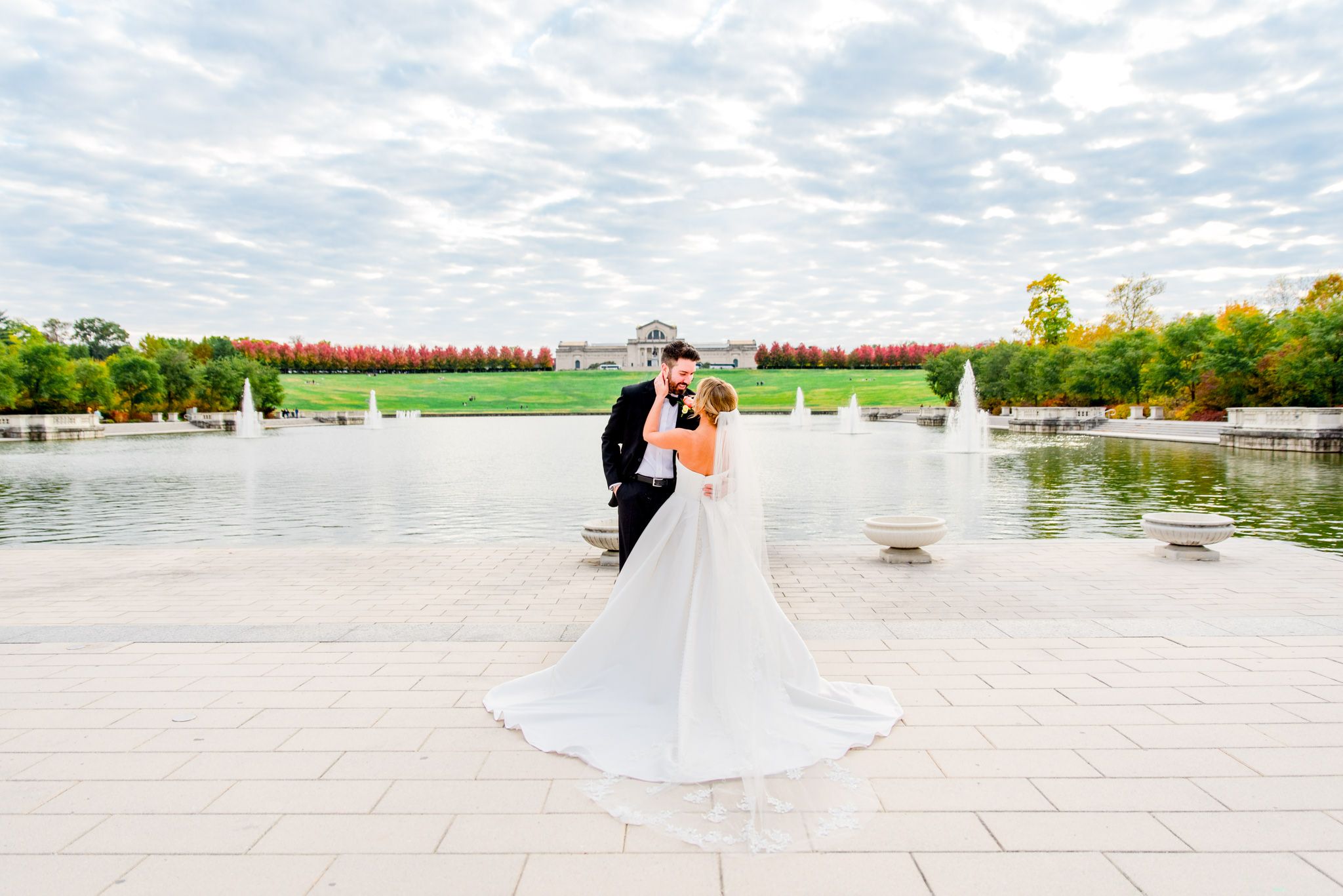 bride's wedding dress and veil fanned out next to groom under blue sky with clouds for Forest Park STL fall wedding