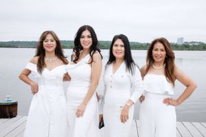 Four women in white outfits pose together on a waterfront dock on an overcast day.