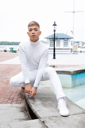 Model in all-white outfit poses casually by marina on brick walkway.