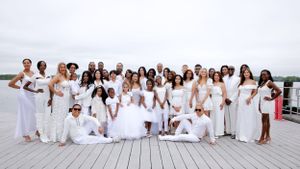 Large group in coordinated white attire poses together on wooden dock by water.