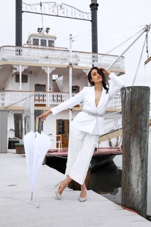 Elegant pose with white umbrella and riverboat in background at waterfront dock.