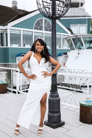 Woman in white dress poses by decorative lamp post with marina and teal building in background.