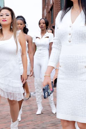 Women in stylish white dresses and outfits pose on brick walkway near building.