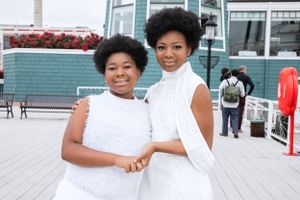 Two people in white outfits pose together on a deck with teal building in background.