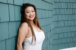 Woman in white tank top smiling against teal shingle wall background.